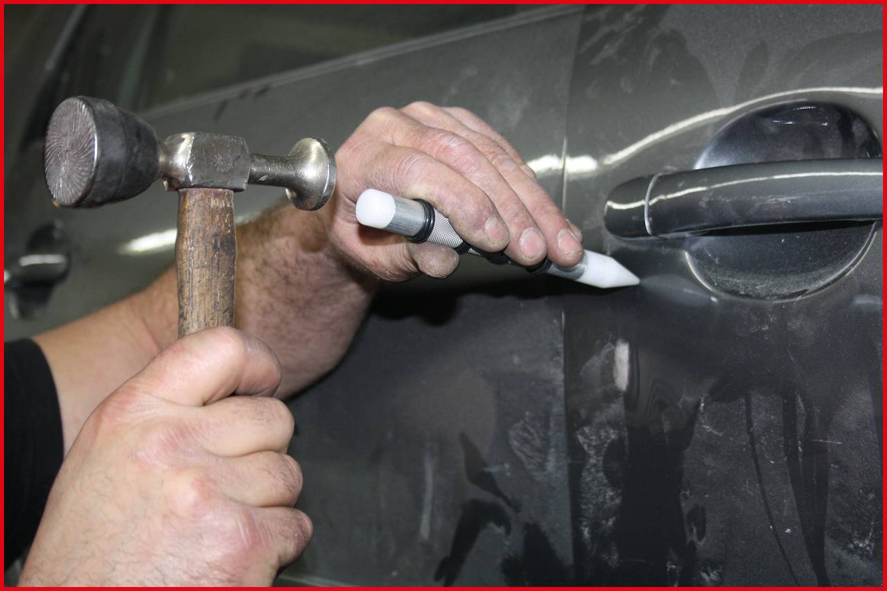 A person is repairing a car door, holding a tool in one hand and a hammer in the other, to remove a dent.