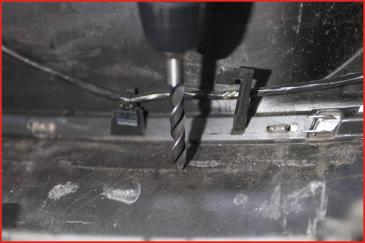 Drill penetrating a metal sheet, close-up. Cables and clips surround the drilling site. The scene depicts a manual work process.