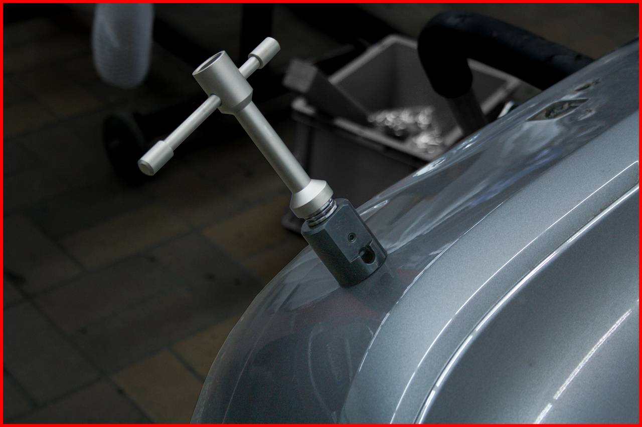 Metal screwdriver secured on a grey car bonnet in a workshop. Blurred details of floor and bucket in the background.