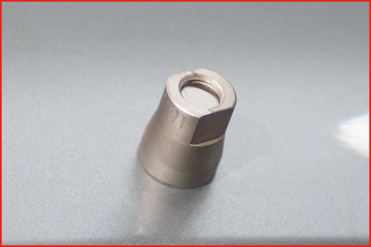 A silver, hexagonal metal component rests on a grey surface. It appears to be a solid, functional element.