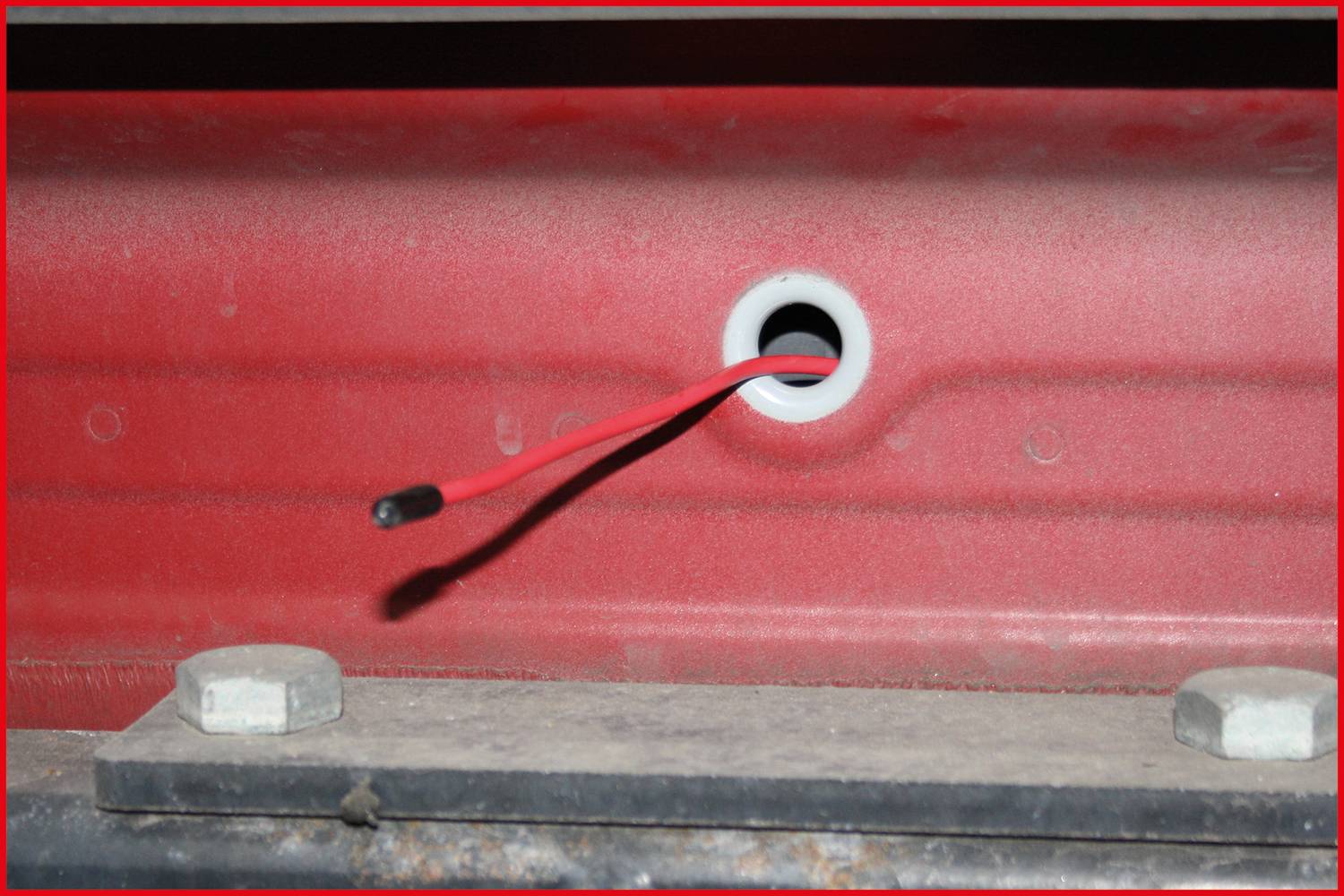 A red cable protrudes from a hole in a red metal surface. There is a mounting bracket beneath it.