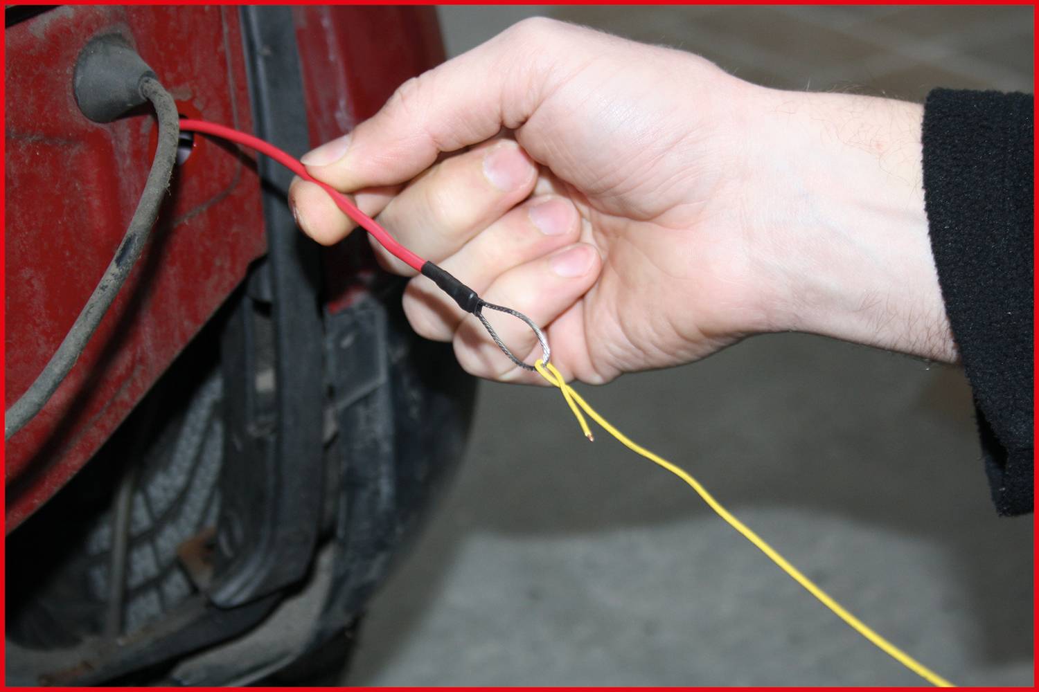 A hand is holding two interconnected wires next to a car tyre. A red and black cable are tied together.
