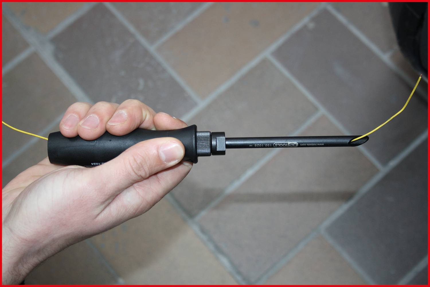 A hand is holding a tool with a black handle and a pointed metal tip. A yellow wire is attached to the tip.