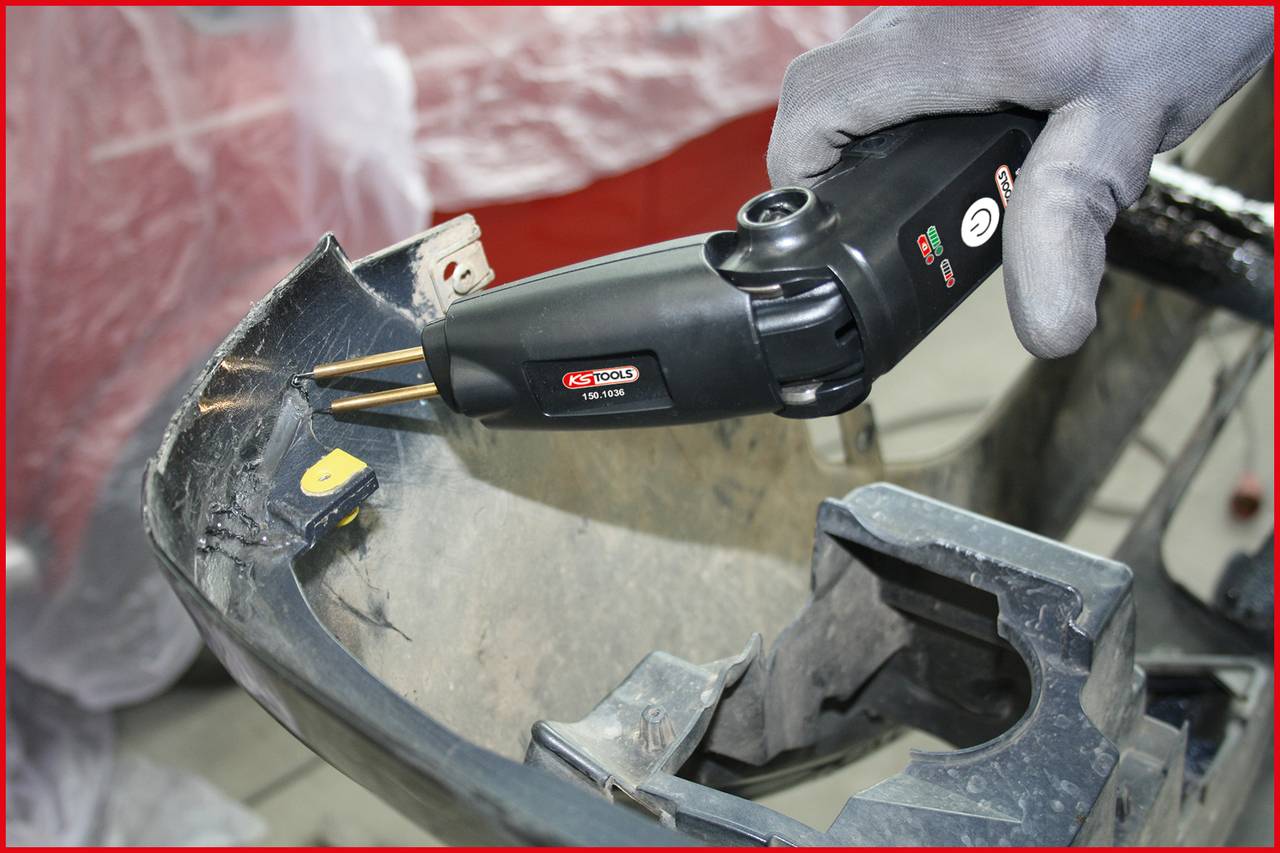A person is repairing a damaged vehicle bumper with a black plastic welding tool in a workshop environment.