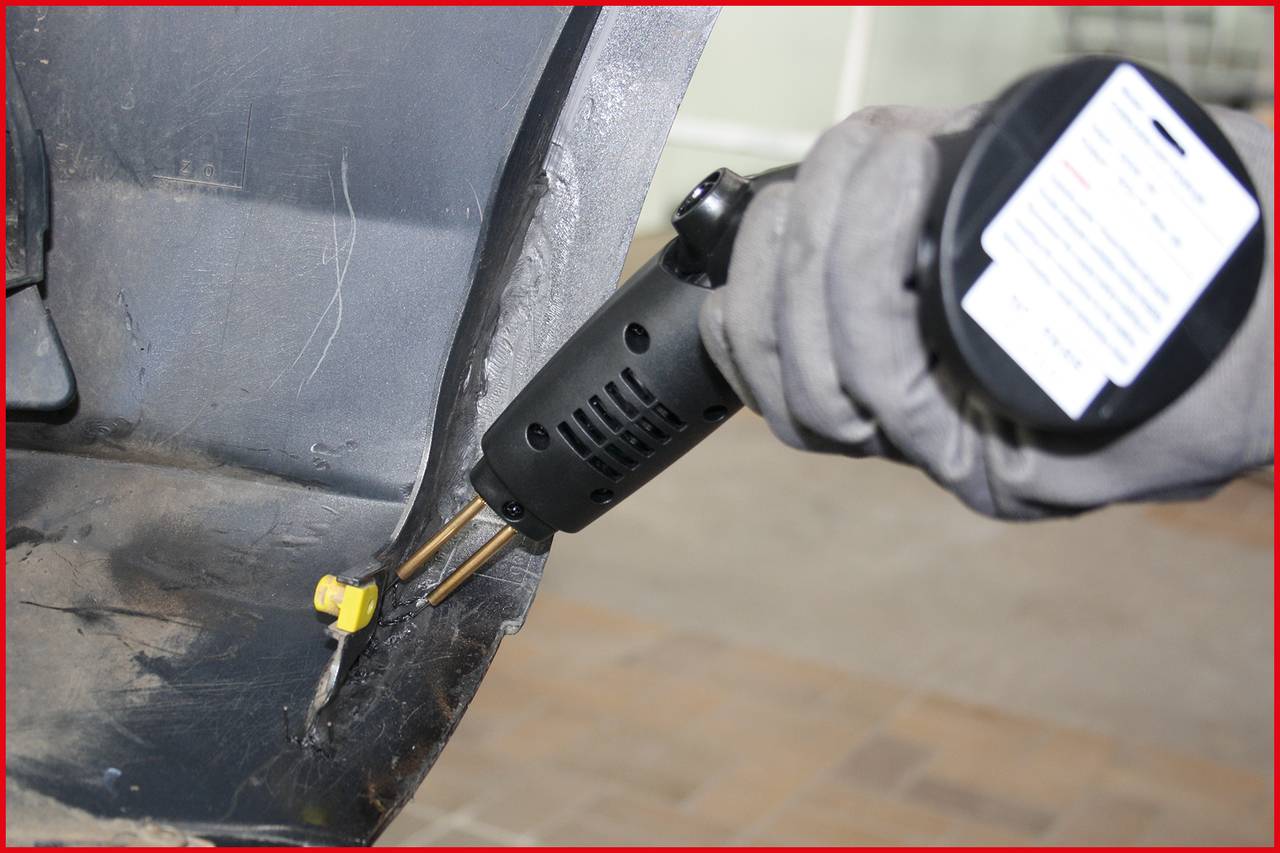 A person is wearing gloves and holding a heating device to repair plastic on a grey vehicle part.