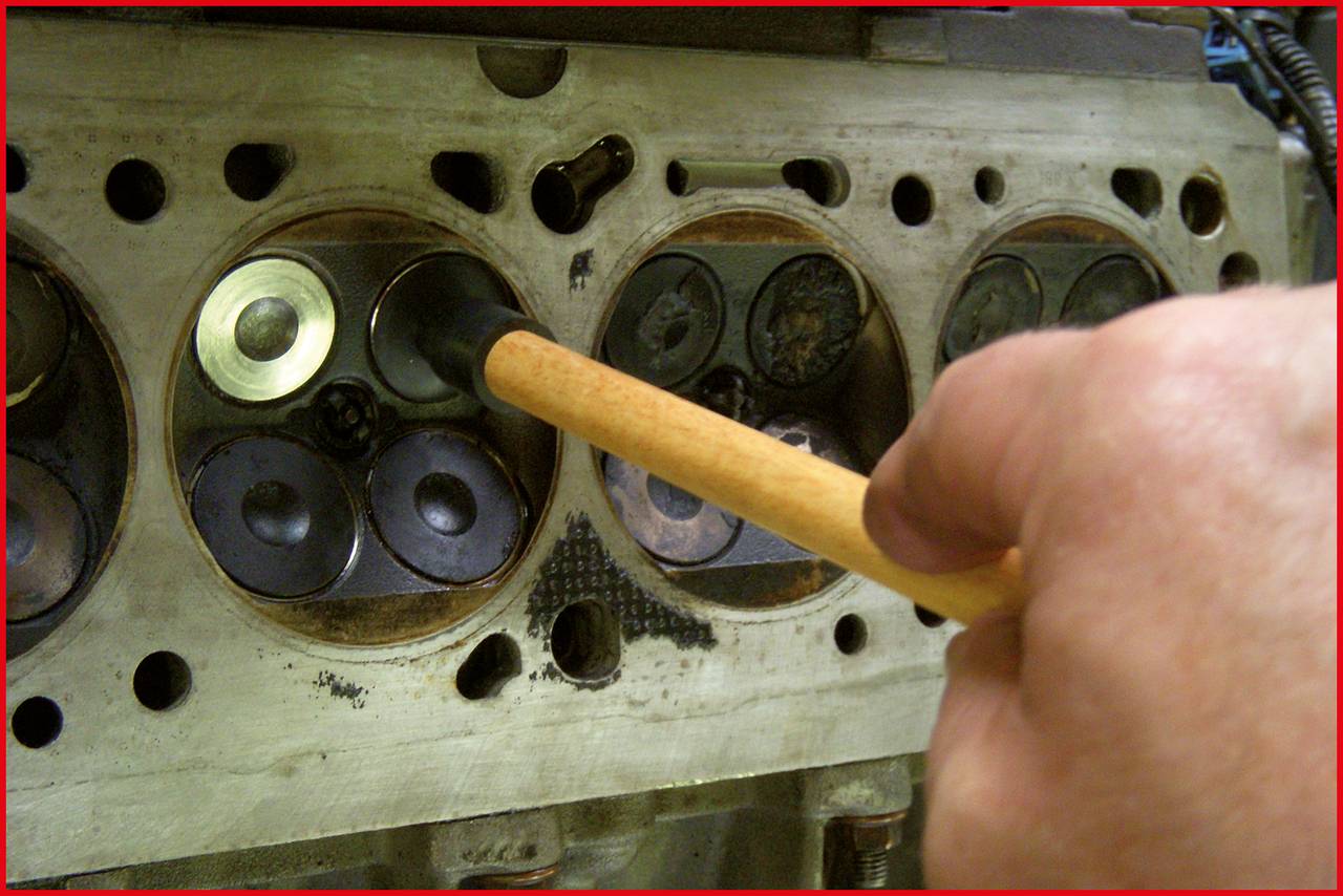 A hand is holding a tool pointing at a cylinder head of an engine, surrounded by valves and metal components in a workshop environment.