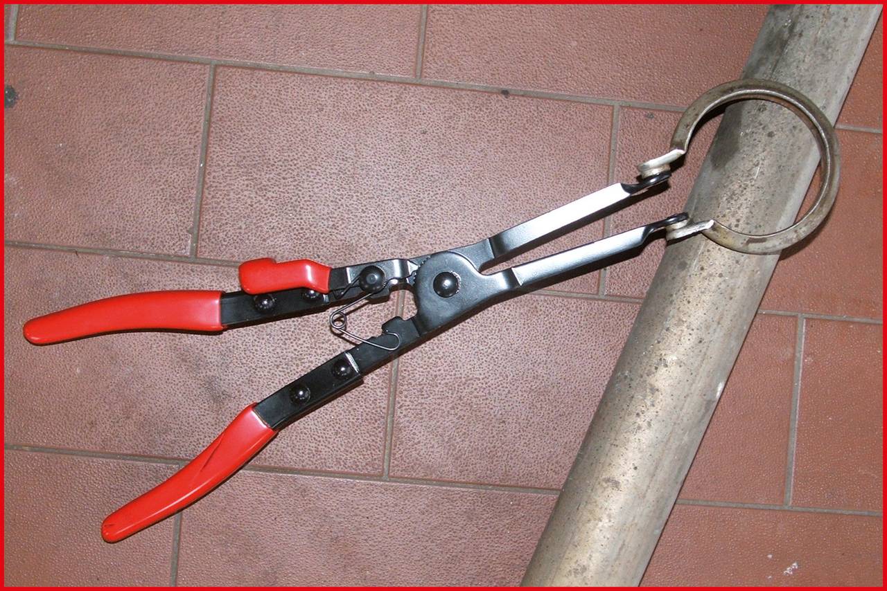 Red pliers attached to a metal rod on a brown tiled floor. Tool with long handles and a narrow gripping tip.