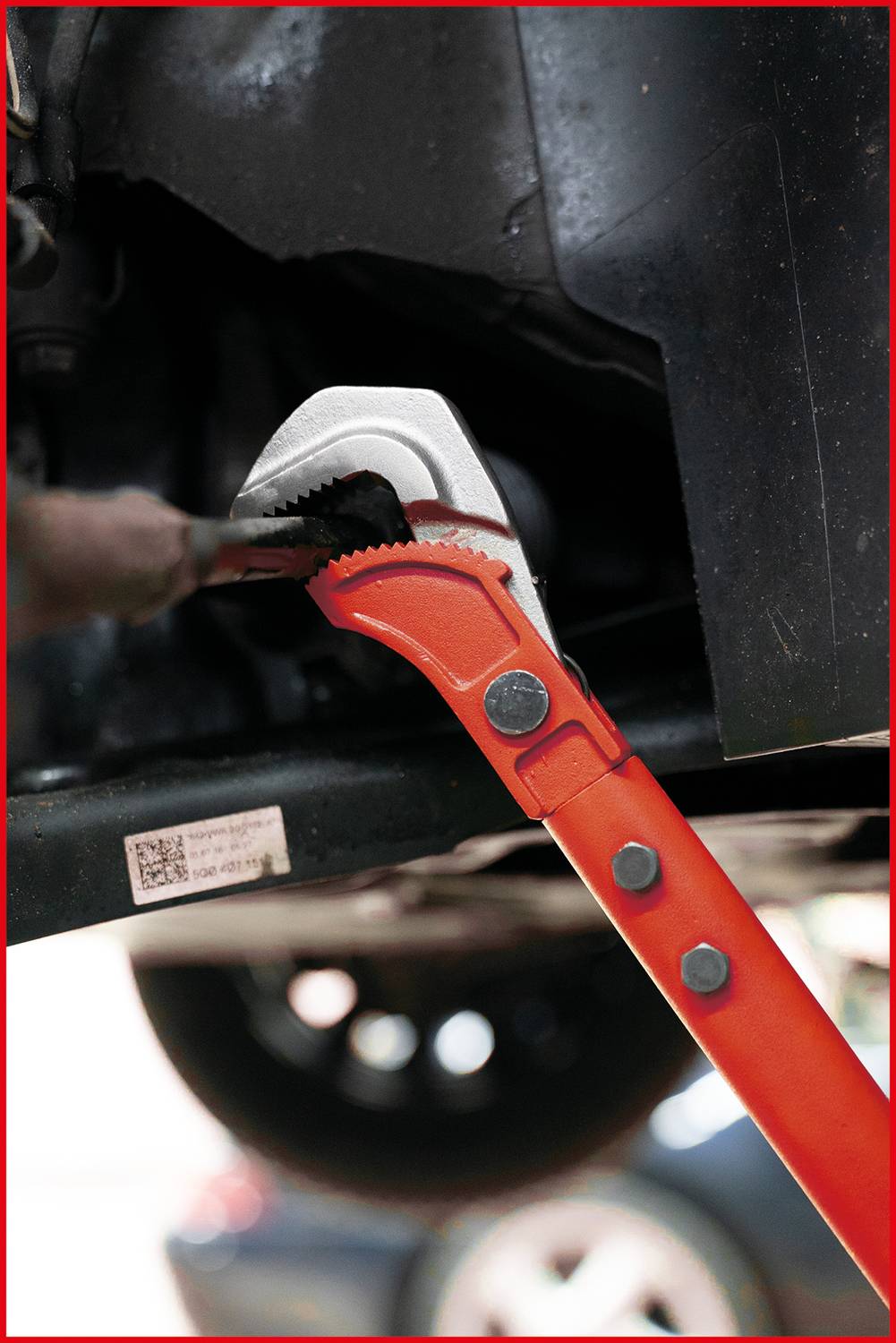 A red adjustable spanner is working on a black metal component underneath a car. In the background, a blurred wheel is visible.