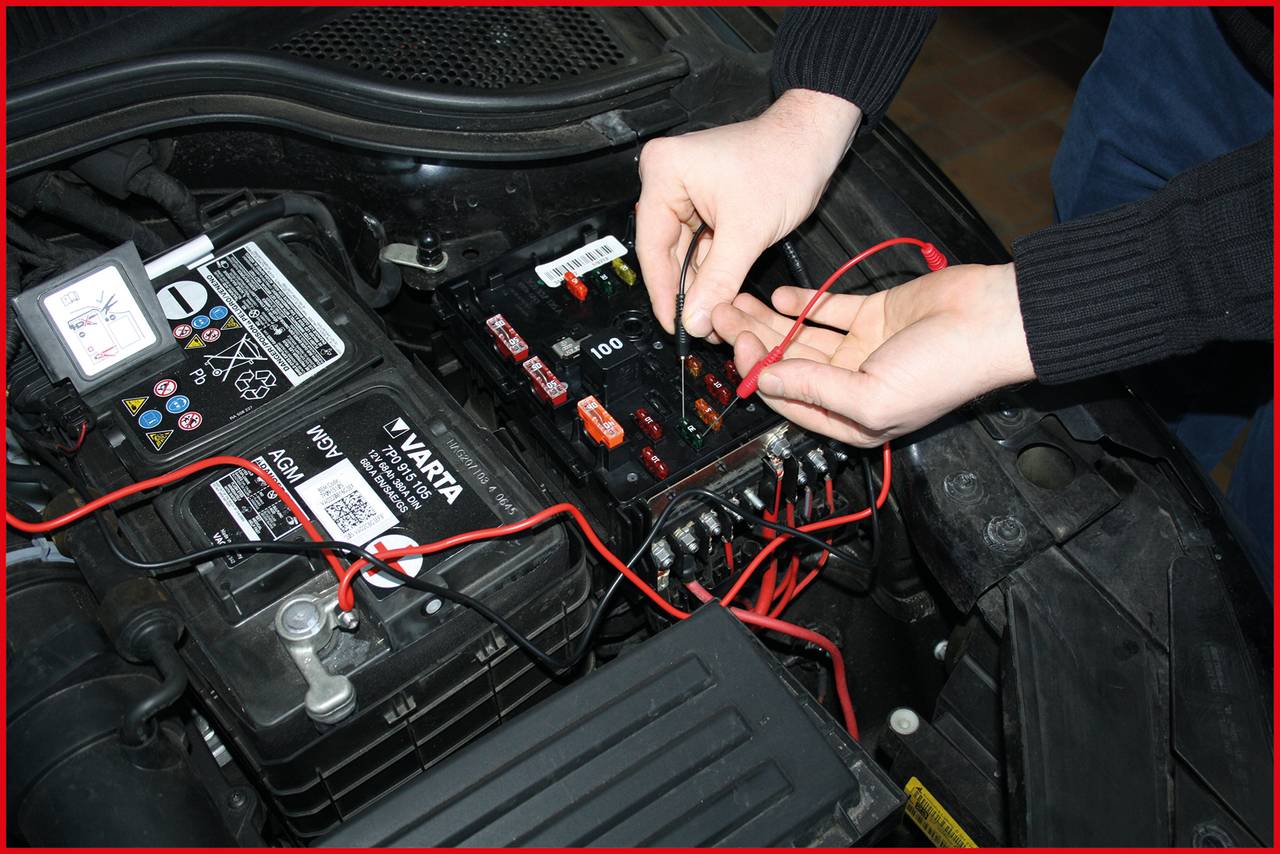 'Two hands working on a car battery with cables and fuses, surrounded by various car parts in the engine compartment.'