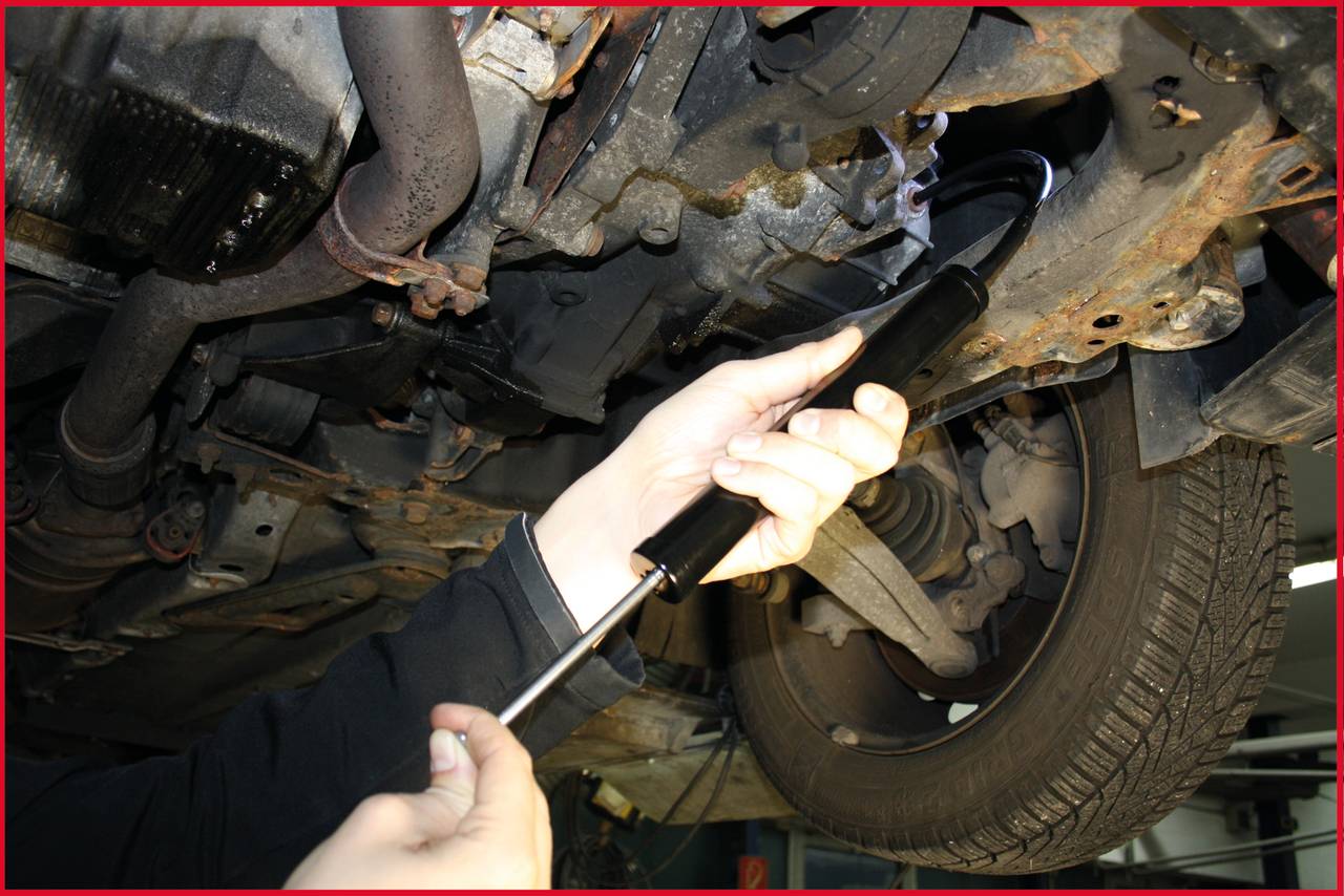 A mechanic is repairing the underside of a car in a workshop. The person is holding a tool and working on the vehicle's suspension.
