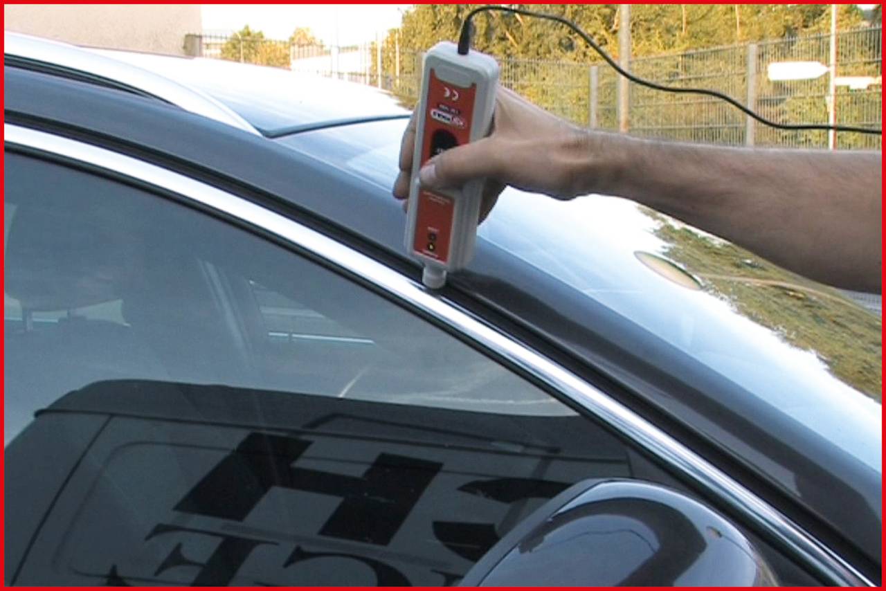 An arm holds a measuring device against the car window. Background slightly blurred, focus on the measurement.