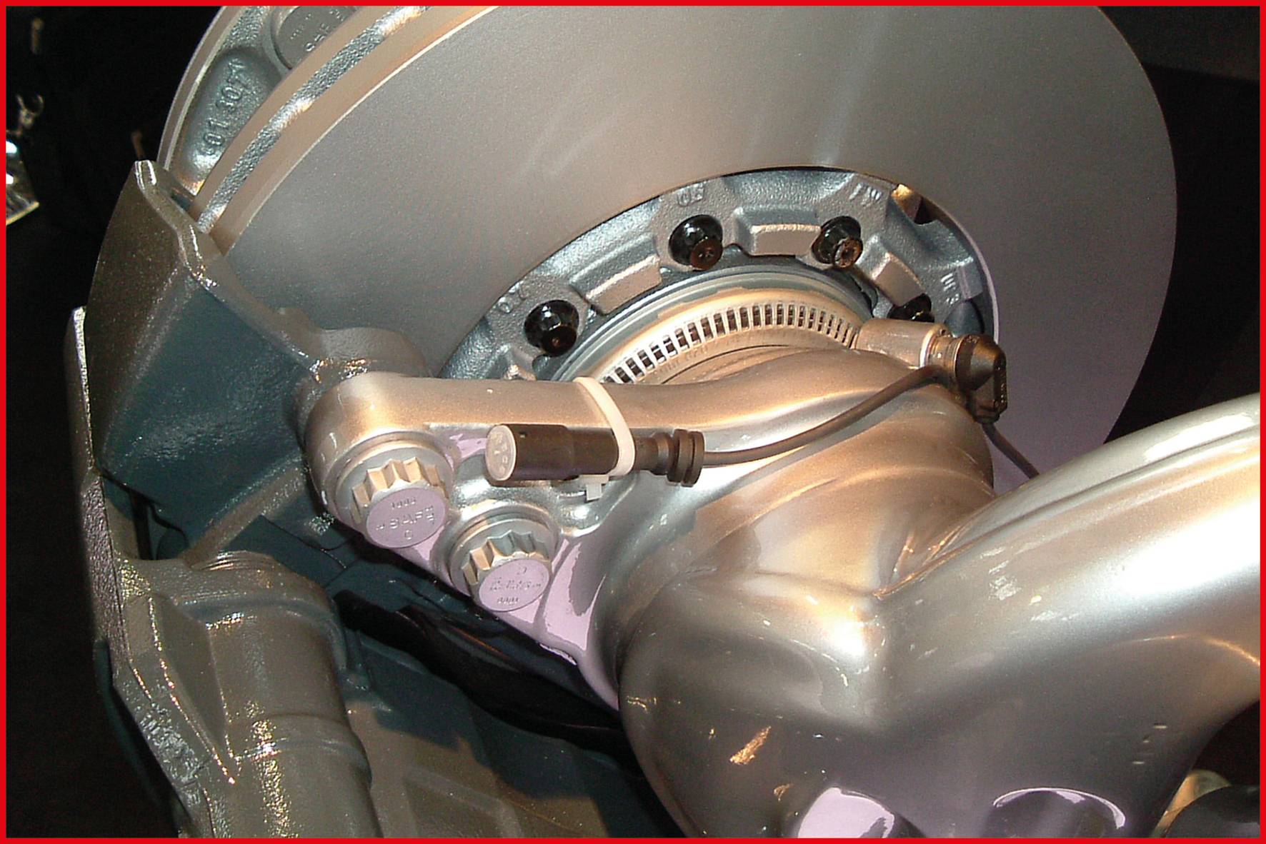 Close-up of a silver car brake disc with brake calliper, cables, and mounting parts in a workshop.