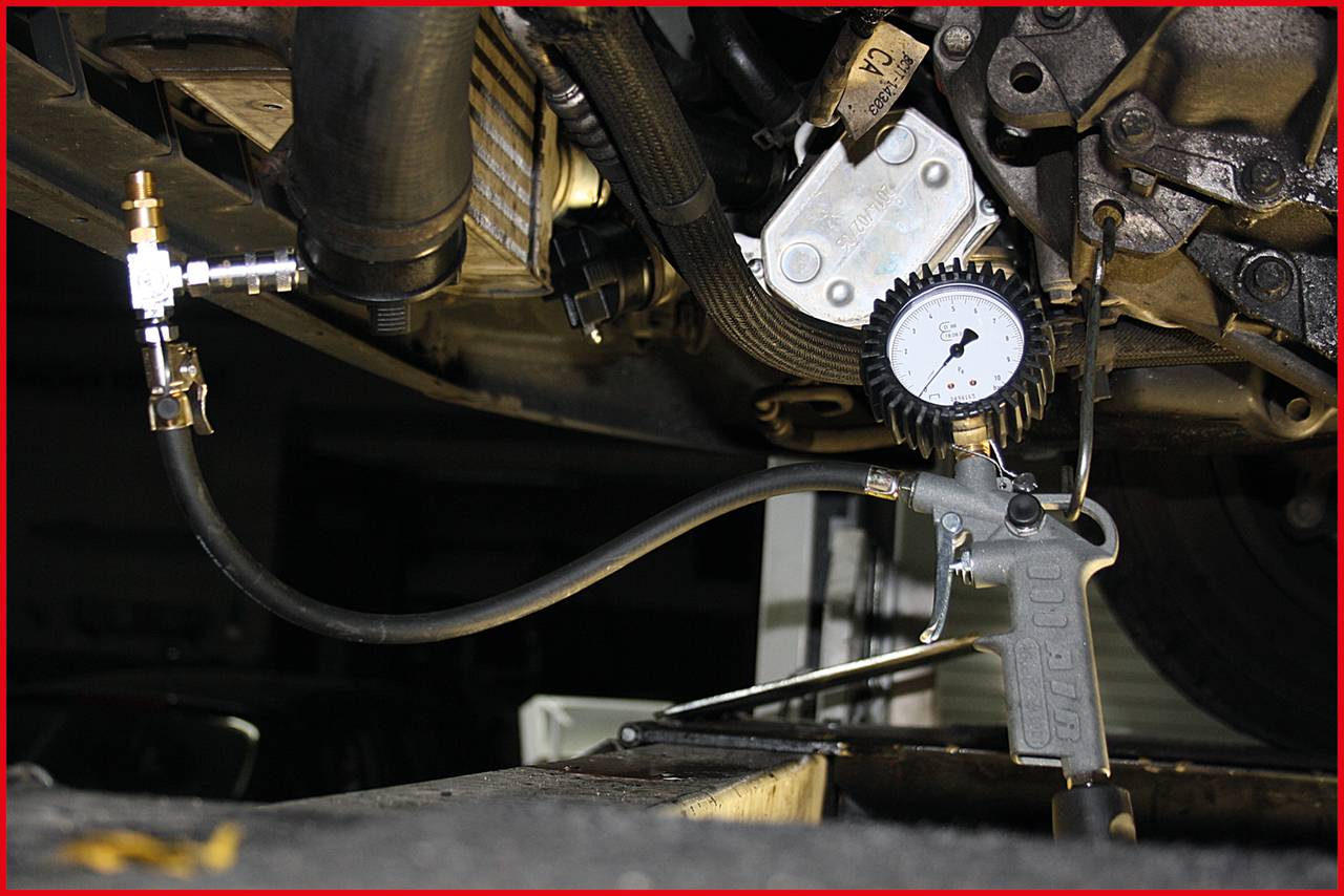 Close-up of a vehicle underside during a pressure test. A pressure gauge is connected to the engine to measure the pressure.