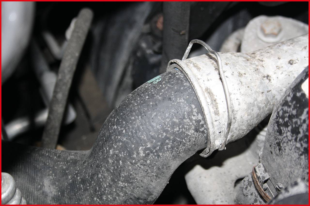A dirty rubber hose with a metal clamp in an engine bay. Dirt deposits are visible on the surface.