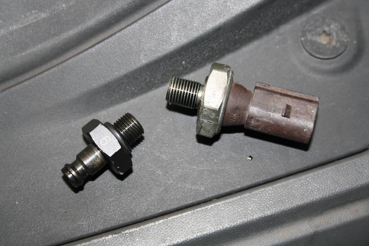 Two oil pressure sensors of different designs are lying on a grey surface. The left one is smaller and cylindrical, the right one larger and conical.