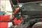 A person wearing red gloves is holding a manometer near an open engine compartment, potentially for pressure measurement.