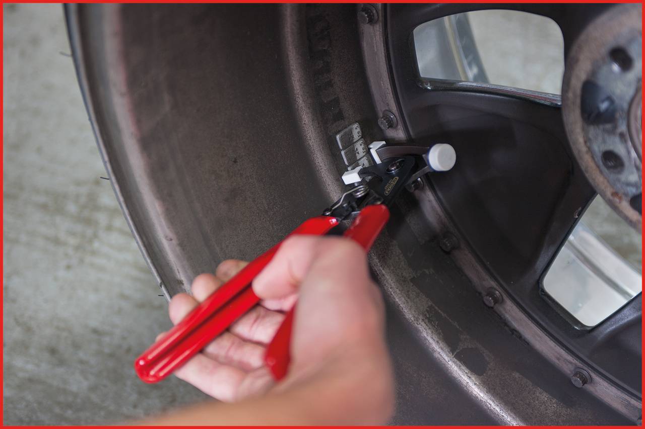 A hand is using a red pliers to attach weights to a vehicle's wheel.