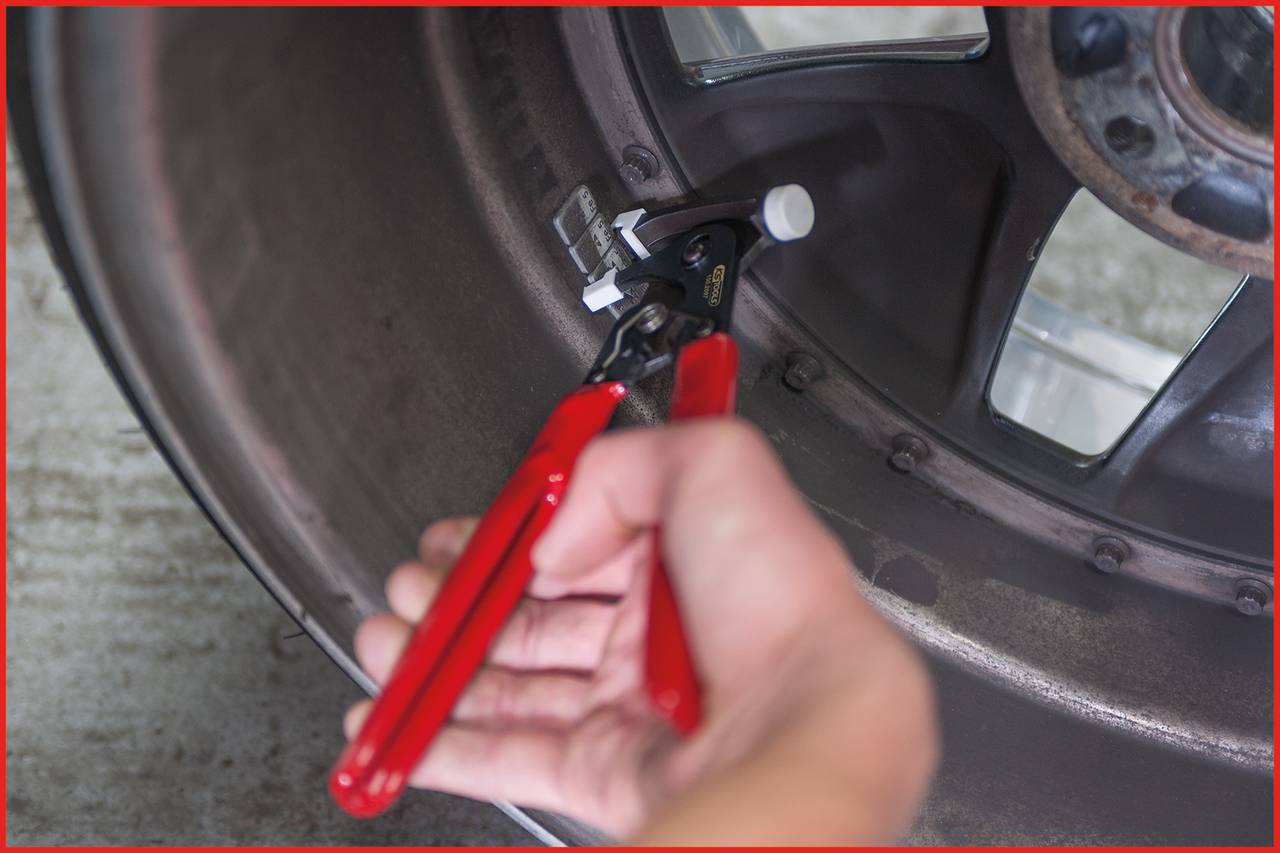 A hand grips a red tool handle, removing a wheel weight from a car tyre rim. A close-up emphasises the process of wheel balancing.