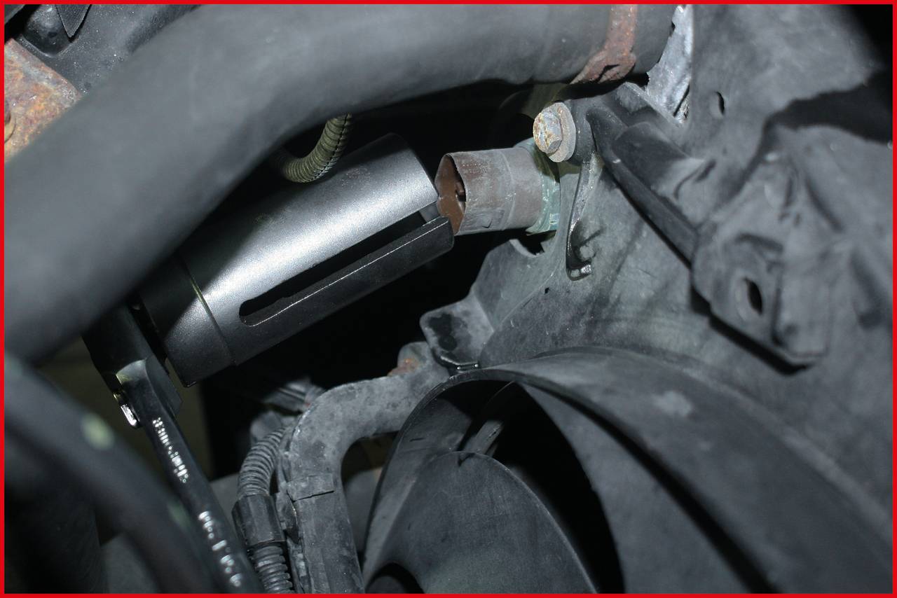 A component in an engine compartment shows a metal cylinder mounted on a bracket, surrounded by hoses and cables.