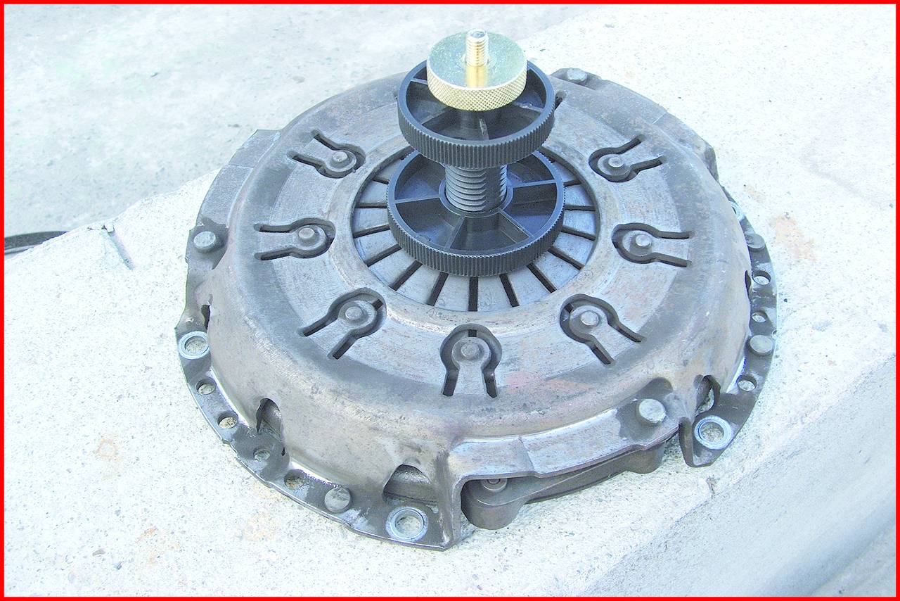 A clutch disc with pressure plate on a concrete surface, central threaded bolt and black, wheel-shaped structure visible.