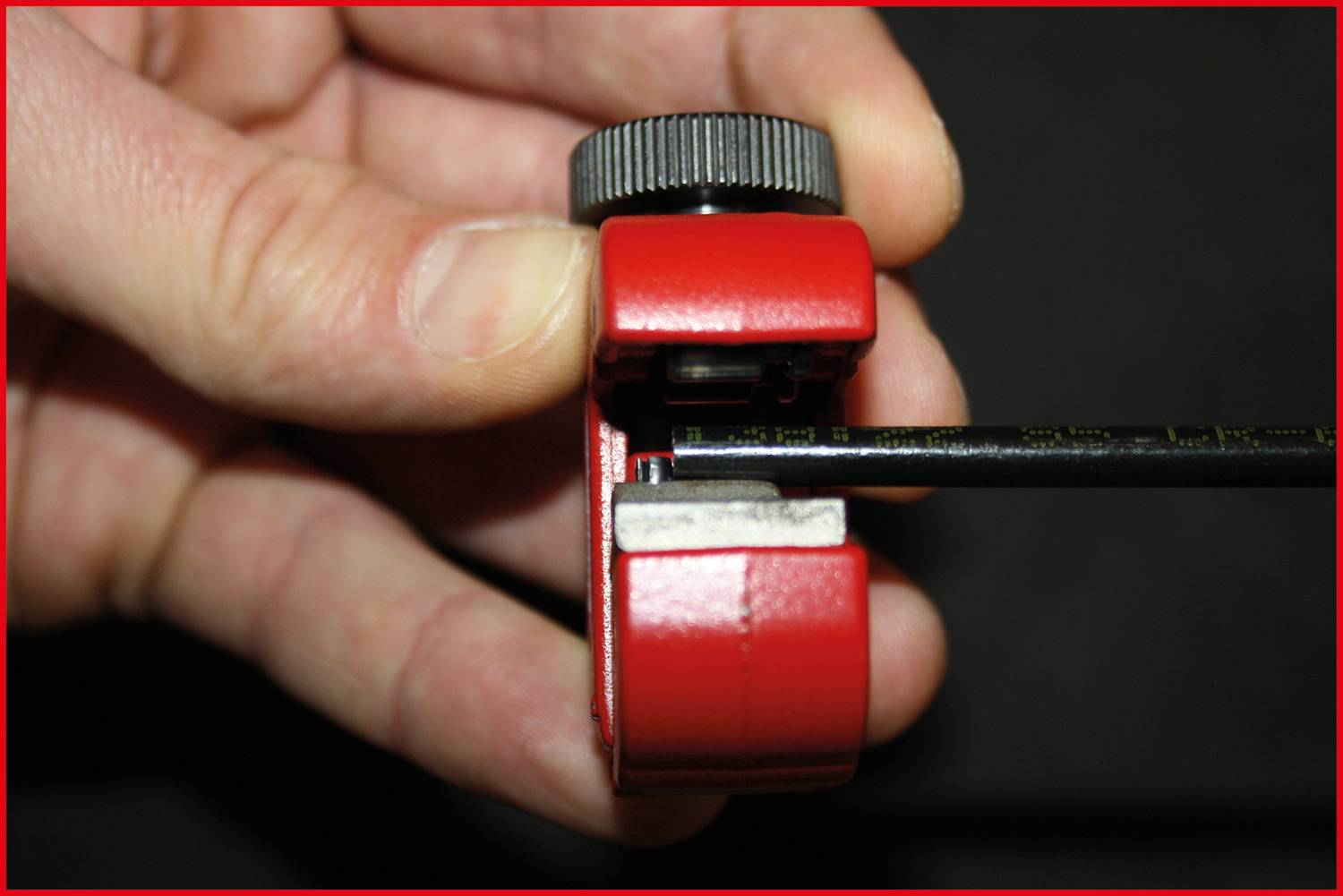 Close-up of a hand holding a red pipe cutter and cutting a pipe. The tool is partially compressed.
