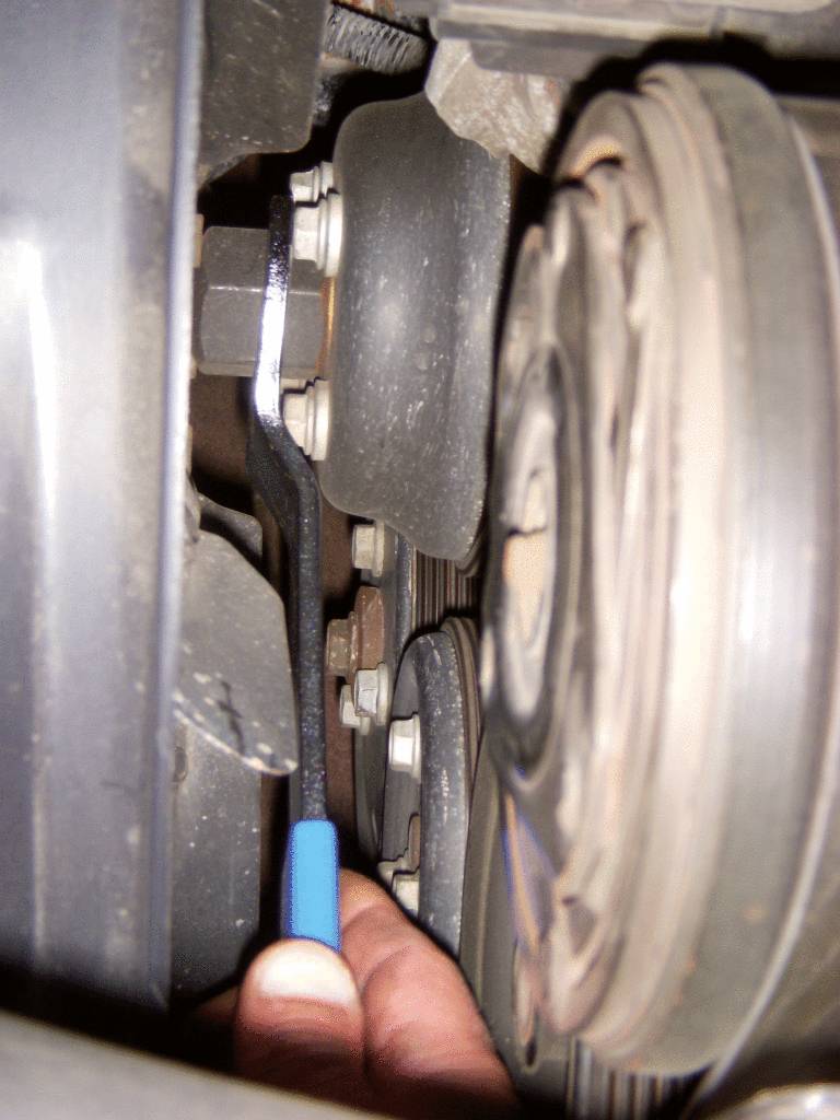 A hand is holding a blue spanner near an automotive part, possibly working on a timing belt or pulley.
