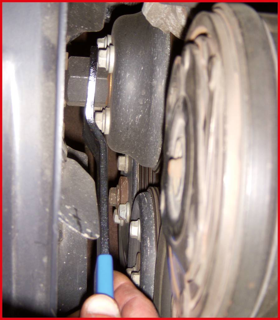 Close-up of a car component: A hand is using a blue tool to inspect the condition of a timing belt between two pulleys.