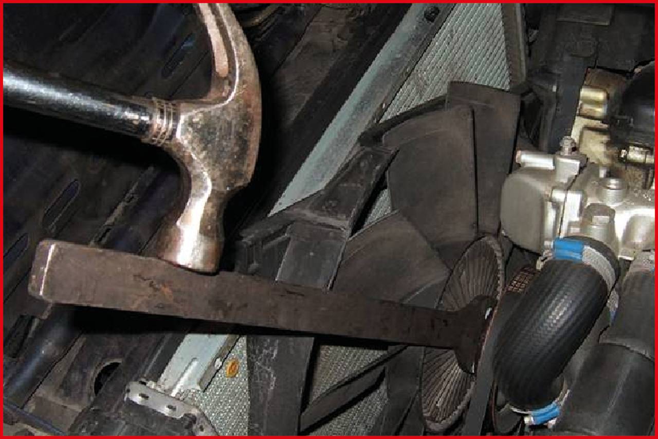 A hammer strikes a metal rod that appears to be fixed to a car radiator. A fan and radiator hoses are visible.