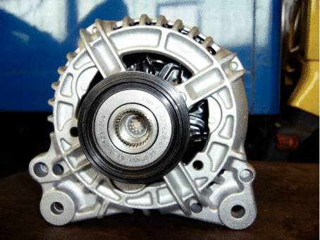 A silver vehicle generator on a table, rear view with visible pulleys and metal ribs.