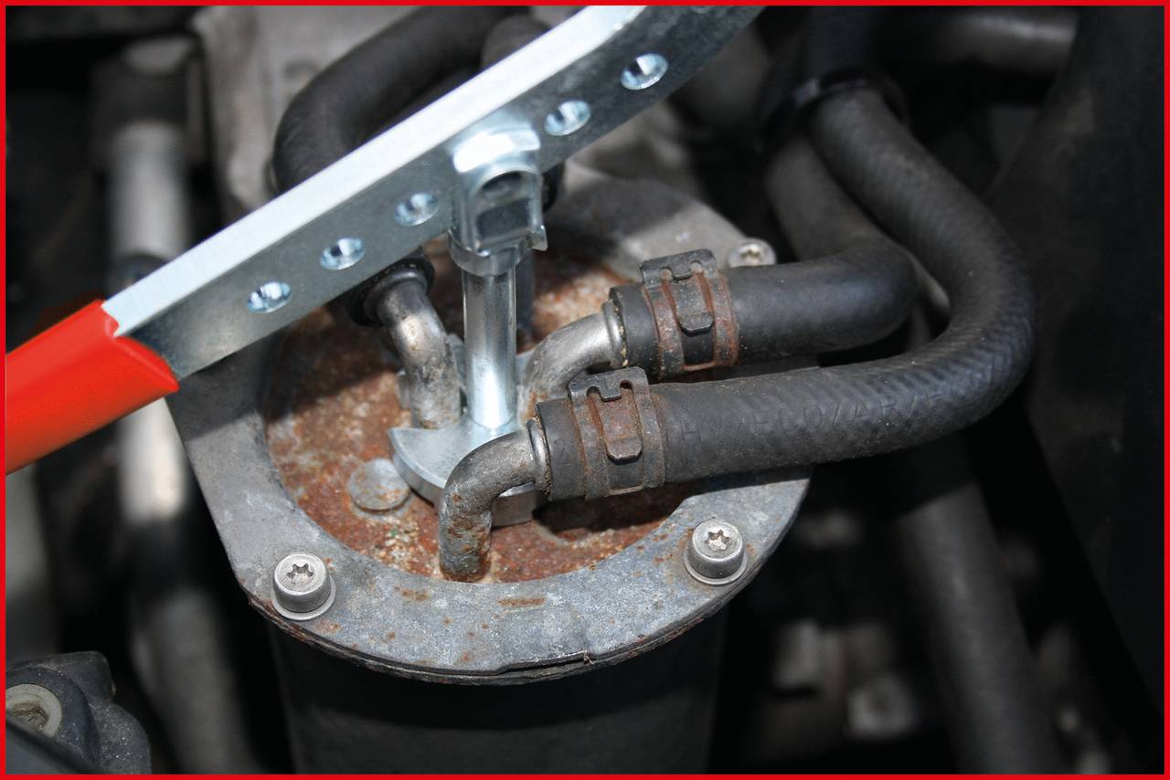 A tool with a red handle removes a rusted metal cover from a car part, likely part of the cooling or heating system.