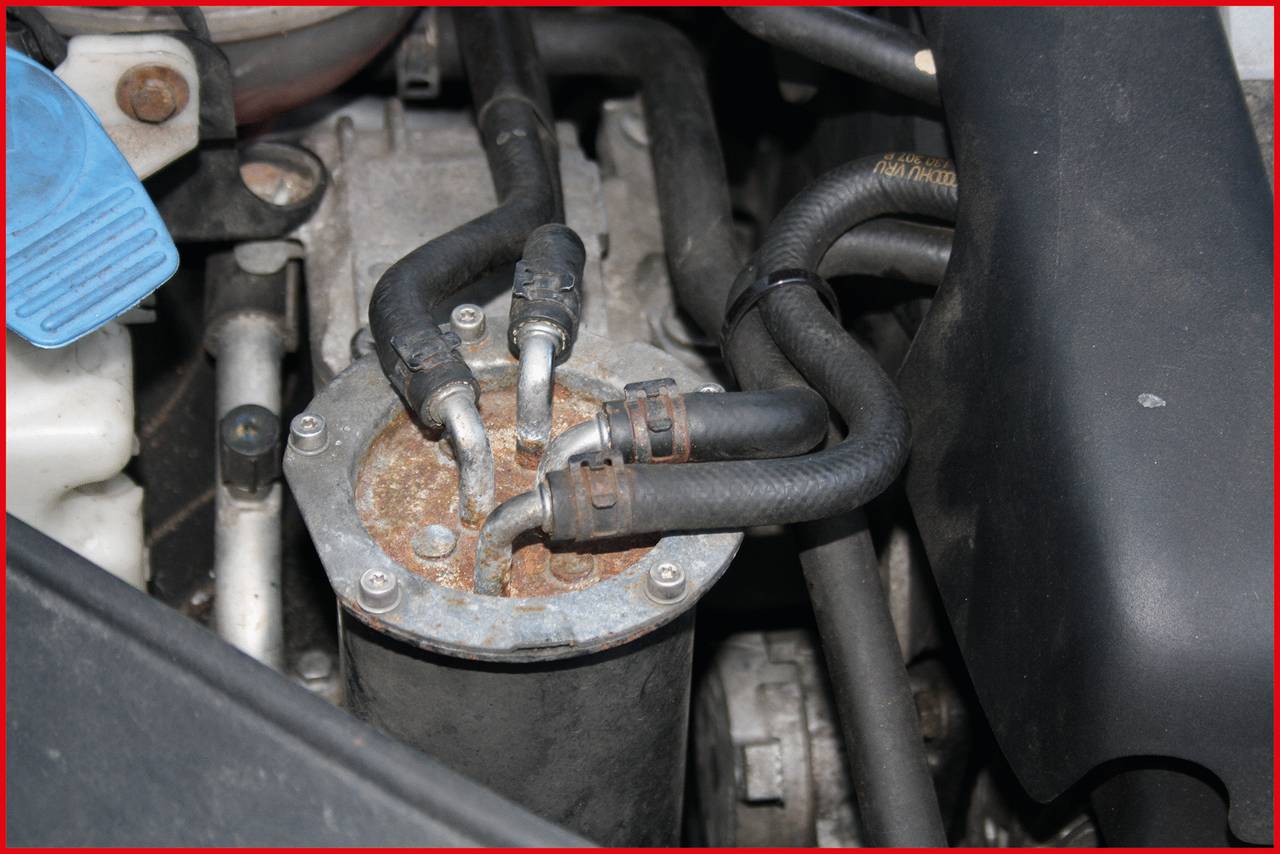 'Old car engine component with rusty connections and hoses situated amongst other engine parts.'