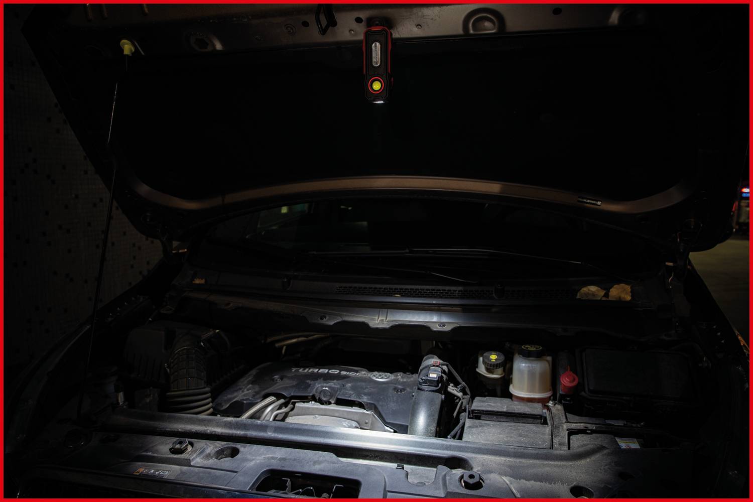 Engine bay of a car with the bonnet open, illuminated by a lamp. The engine and several components are visible.