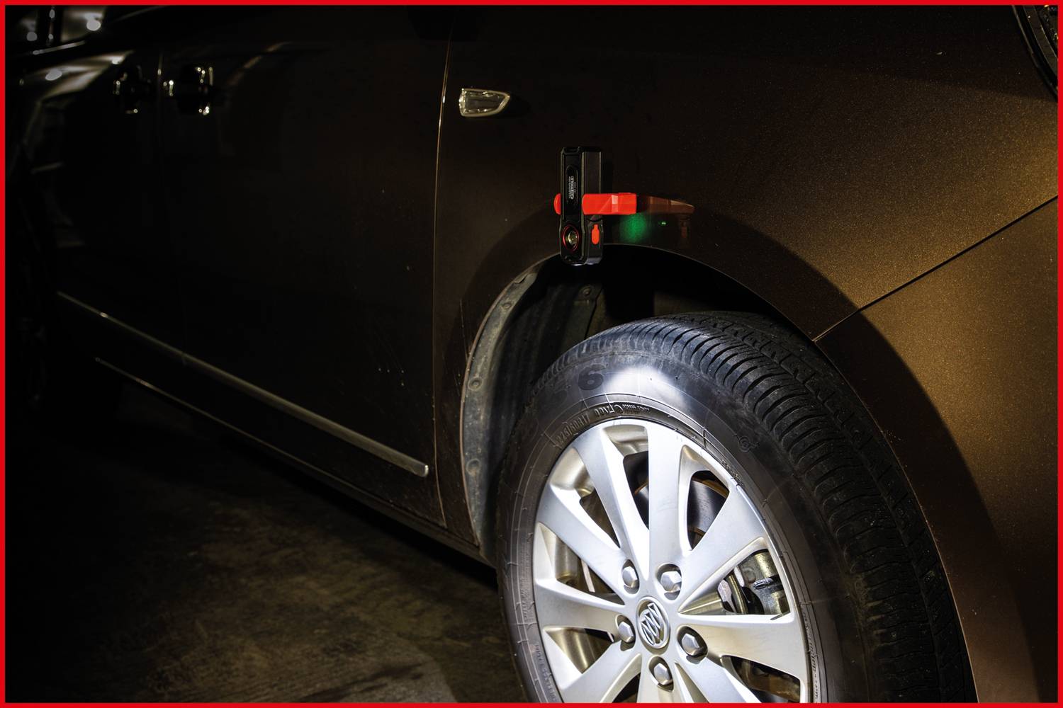 A brown car in the darkness. The focus is on the front wheel, with its tyre illuminated. A small device on the wheel arch.