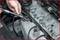 A hand is holding a spark plug over an engine block. The engine shows several removed spark plug locations. Work is being carried out on a car.
