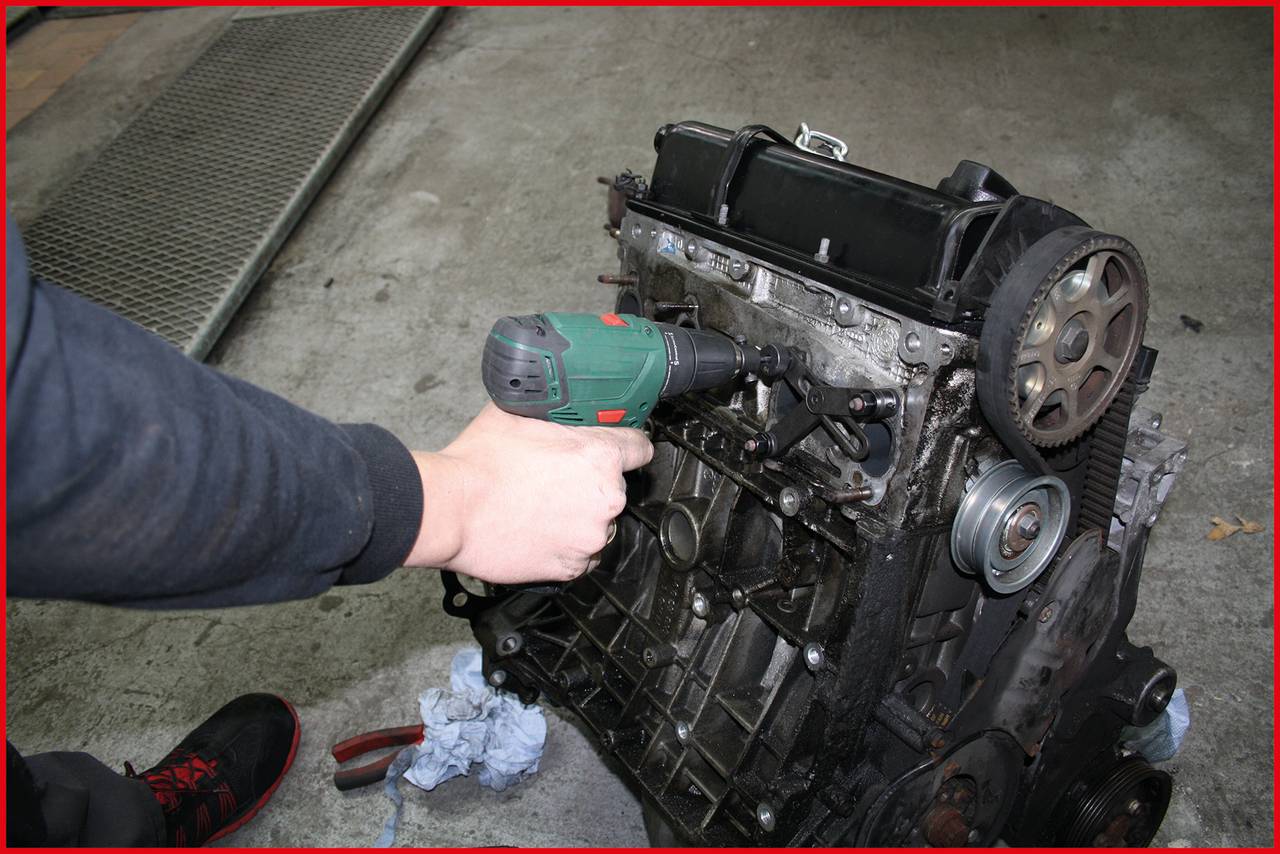 A person is repairing a car engine with an electric power drill in a workshop environment. Tools are lying nearby.
