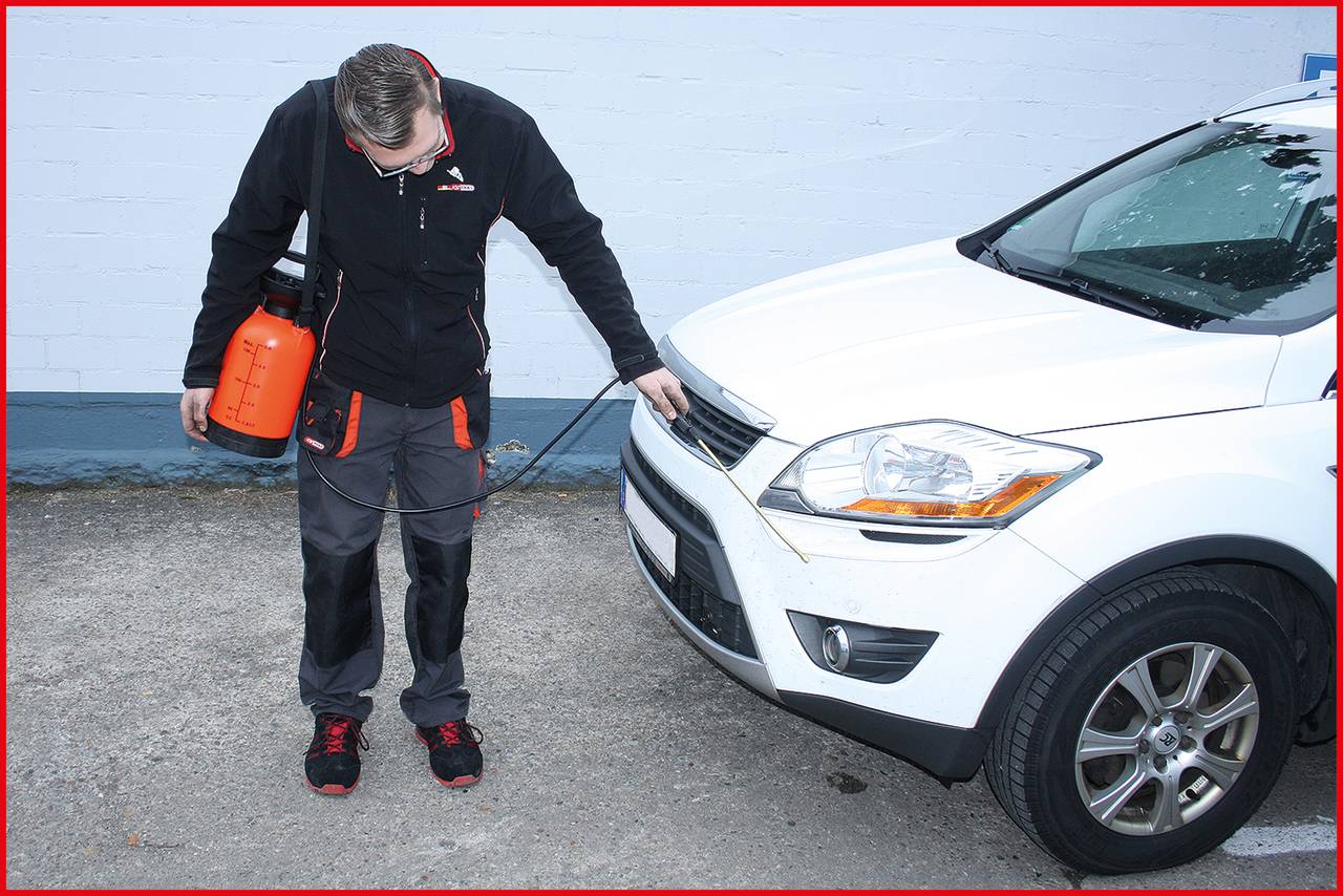 A person cleaning the front of a white car with a spray device, used for vehicle maintenance outdoors.