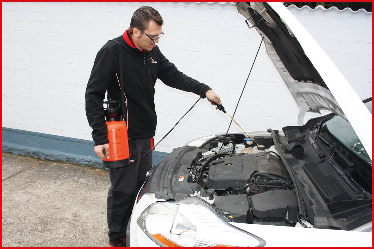 A man is checking the engine of a car while holding a cleaning device in his hand. The bonnet is open.