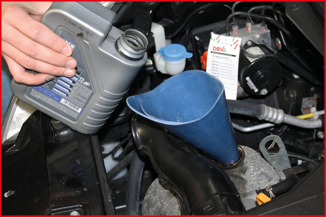 A person is pouring motor oil into a blue funnel in the engine compartment of a vehicle. Several automotive parts are visible in the background.