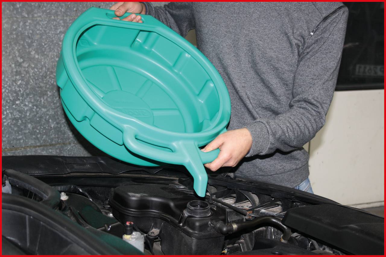 A person is holding a green oil pan over a car's engine bay, ready for an oil change.