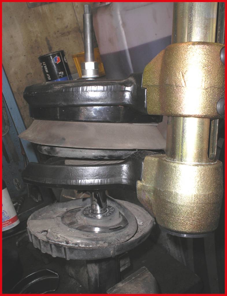 Vehicle spring is being compressed by a hydraulic machine. Workshop environment with tools visible in the background.