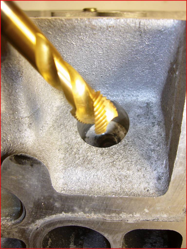 Close-up of a golden twist drill bit machining a metal component. The drill is in operation, cutting a hole or thread.