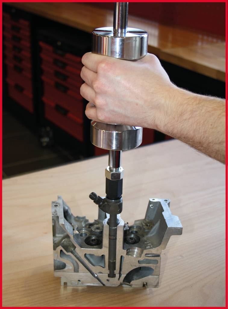 A metal tool is being held by a hand and inserted into a silver motor component. The background shows a workshop.