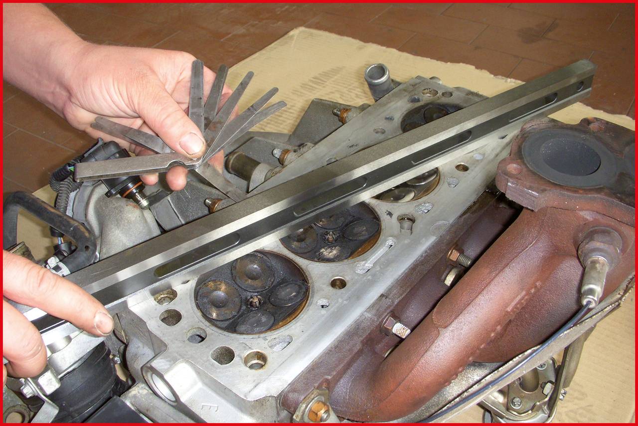 A person is measuring the valve clearance of a cylinder head using a feeler gauge and a straight edge during engine repairs.