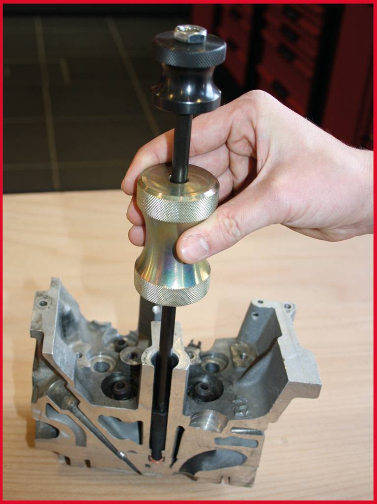 A hand is using a tool to install or remove a valve in an engine block. The engine block is partially disassembled.