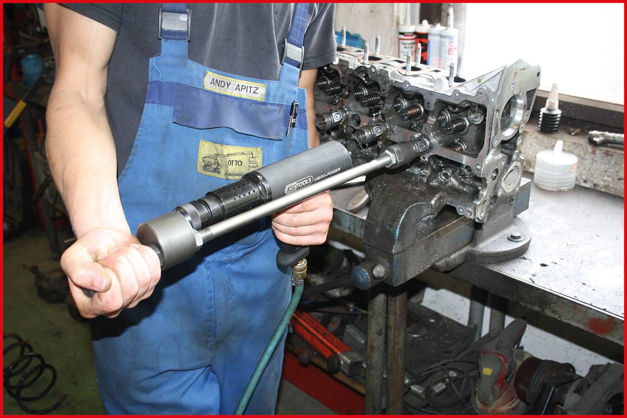 'Mechanic in blue dungarees repairs cylinder head in workshop, holding a pneumatic tool.'