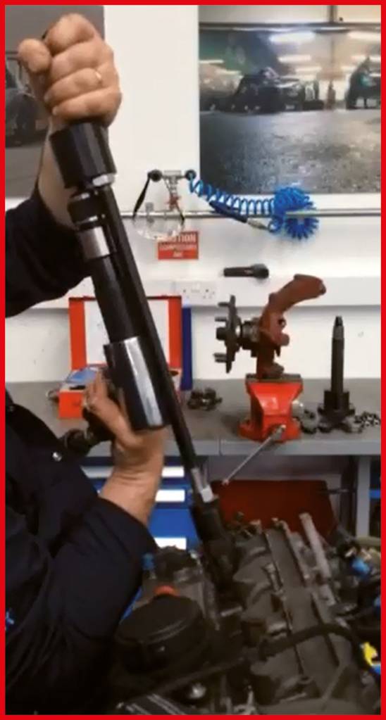 A person is installing a tool on an engine in a workshop environment, surrounded by tools and automotive parts.