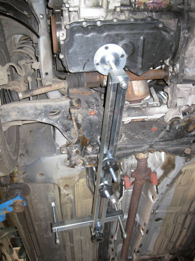 A car is being repaired on a vehicle lift. An engine puller is attached to the engine block, which is visible from underneath.