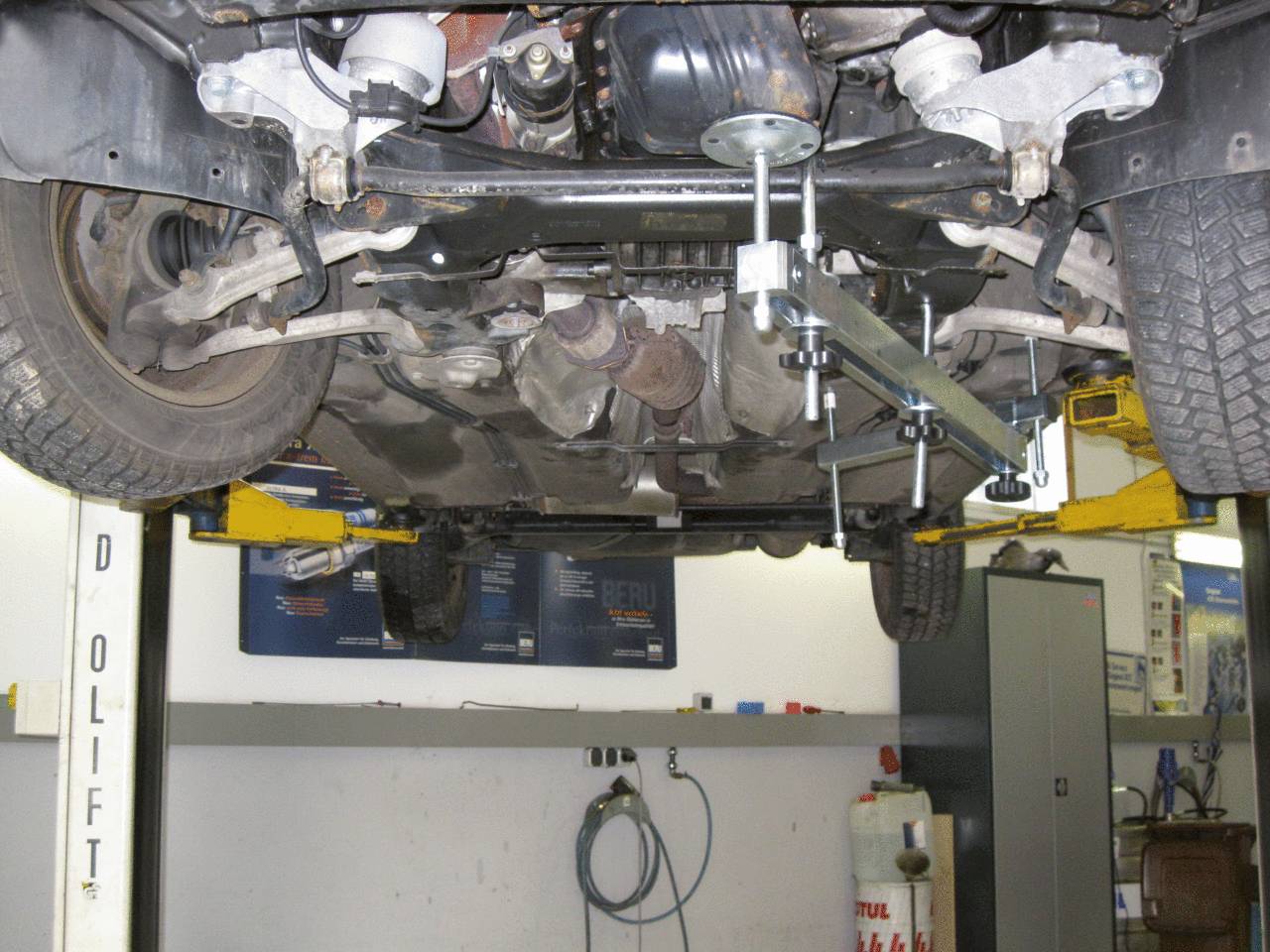 Car on a lift in a workshop, underside and exhaust visible. Mechanical work in progress. Tools and cabinets in the background.