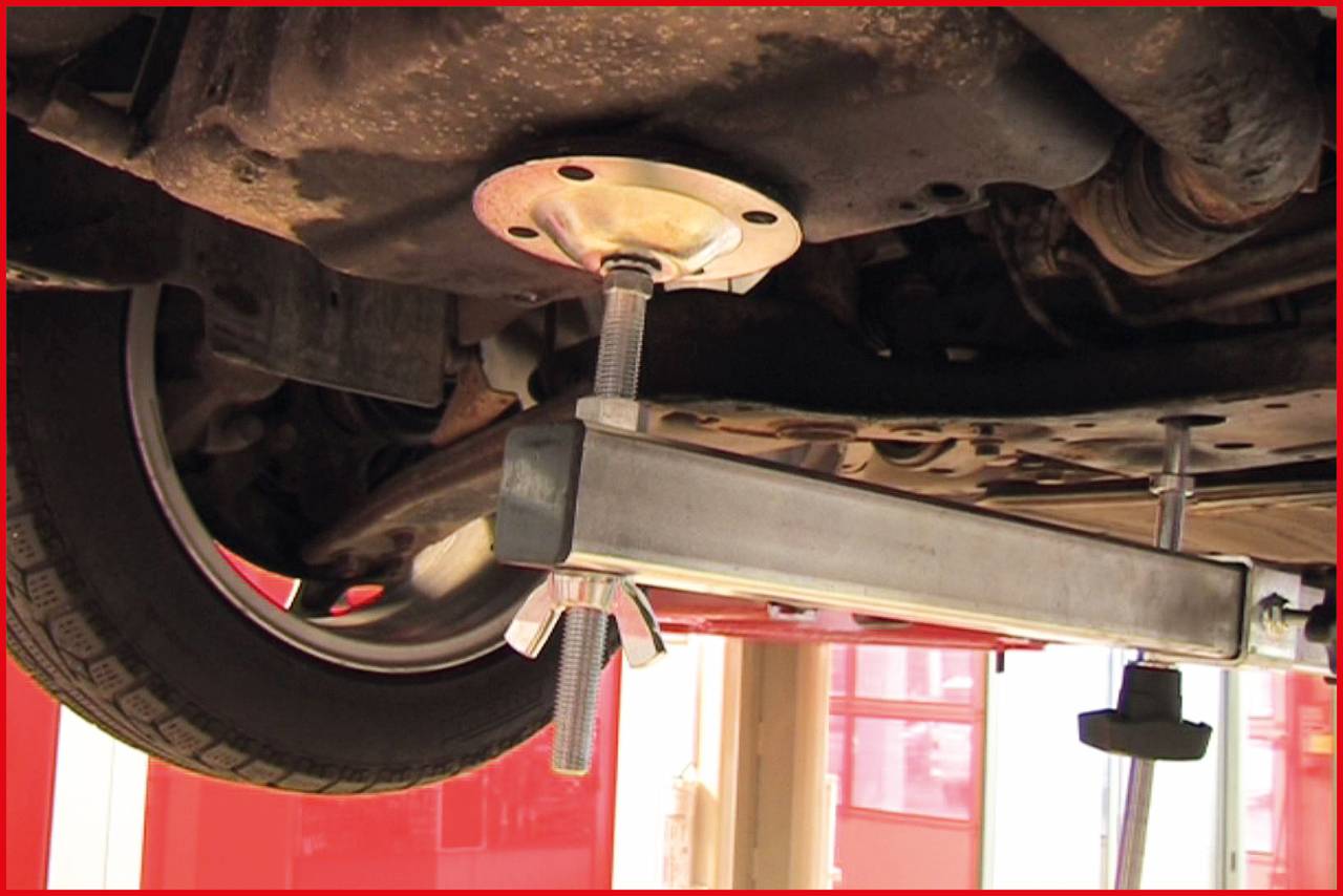 A car tyre is being lifted by a hydraulic lifting device in a workshop. The focus is on the lifting equipment beneath the vehicle.