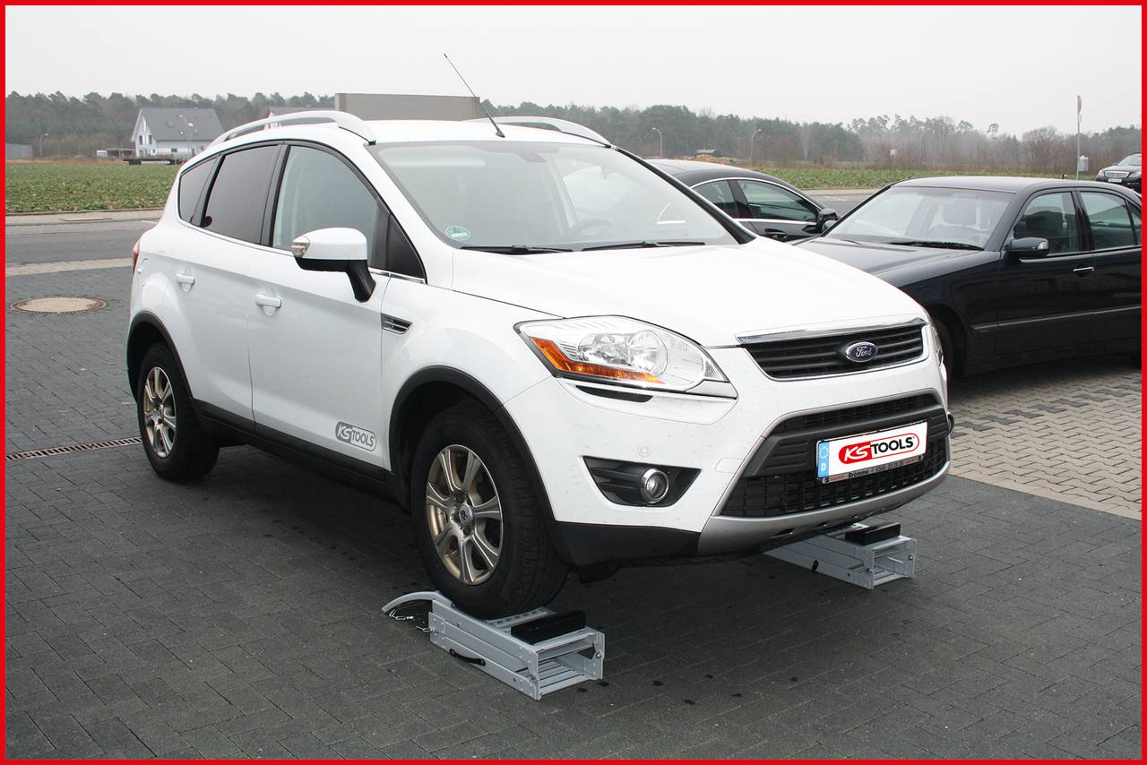 A white car is parked on a mobile tyre-changing assistant, surrounded by another vehicle and a rural road in the background.
